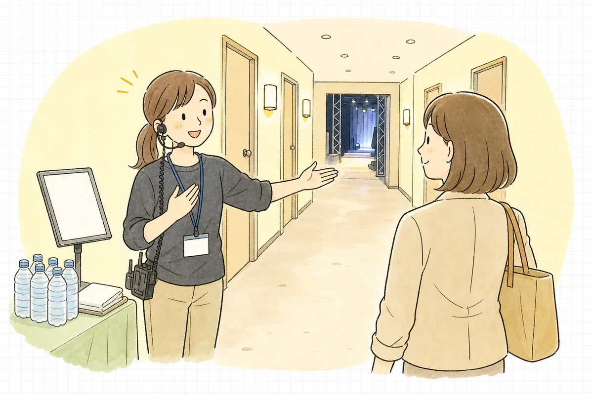 開演前のイベント会場の楽屋前の廊下で、名前を書いた控室の案内を確認しながら、登壇者を笑顔で迎え入れているアテンド担当の制作スタッフ